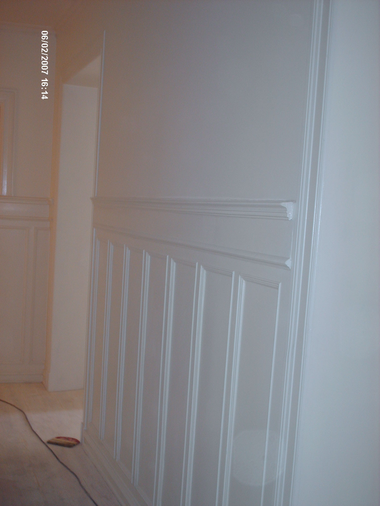 Świeżo pomalowana, jasna ściana z dekoracyjnymi panelami pionowymi i listwami, widoczna podłoga zabezpieczona folią malarską, w tle fragment mebla w podobnym stylu.