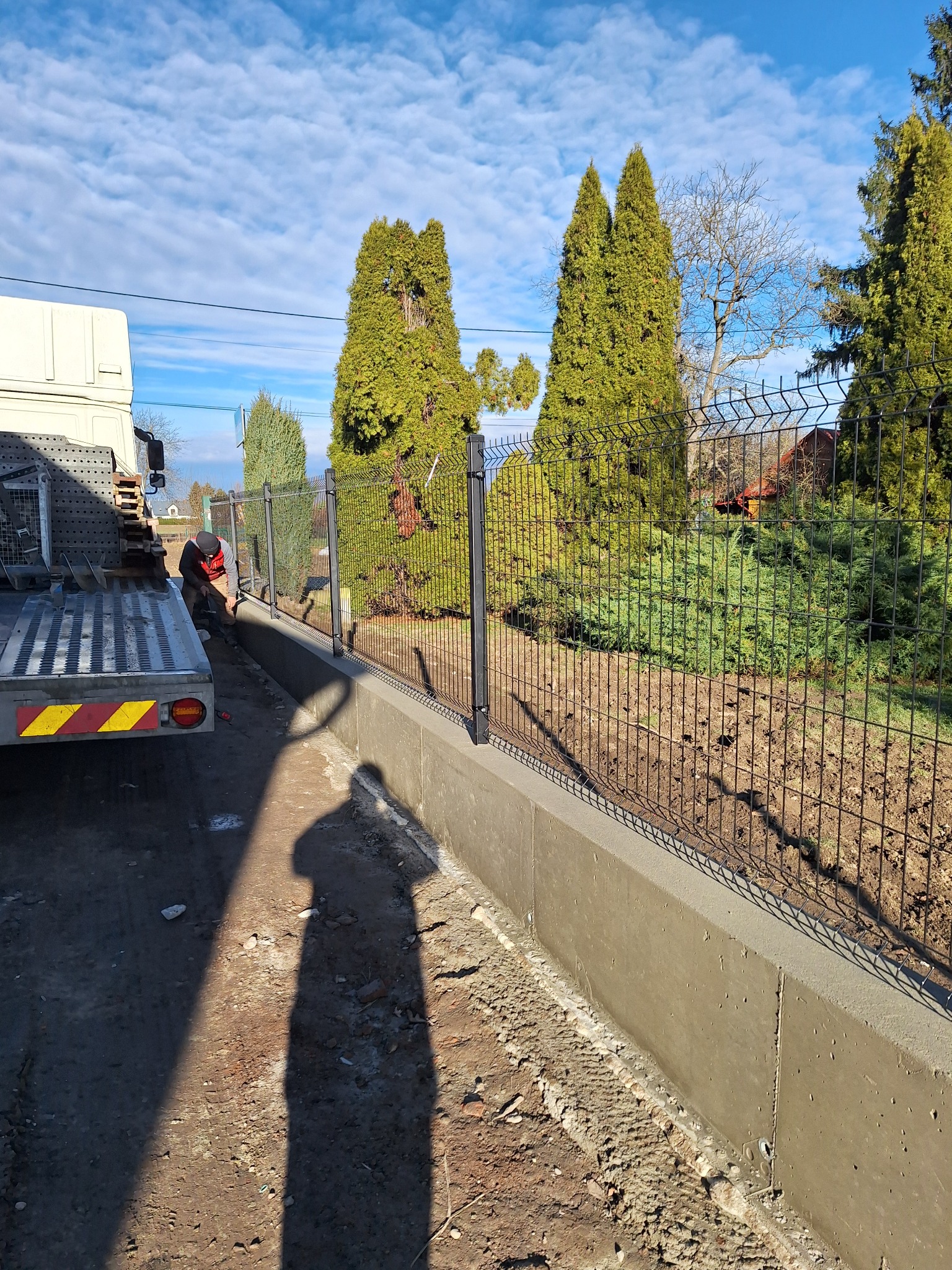 Montaż czarnego ogrodzenia panelowego na betonowej podmurówce w Gródku nad Dunajcem, widoczny pracownik przy ciężarówce i tło z tujami pod błękitnym niebem.