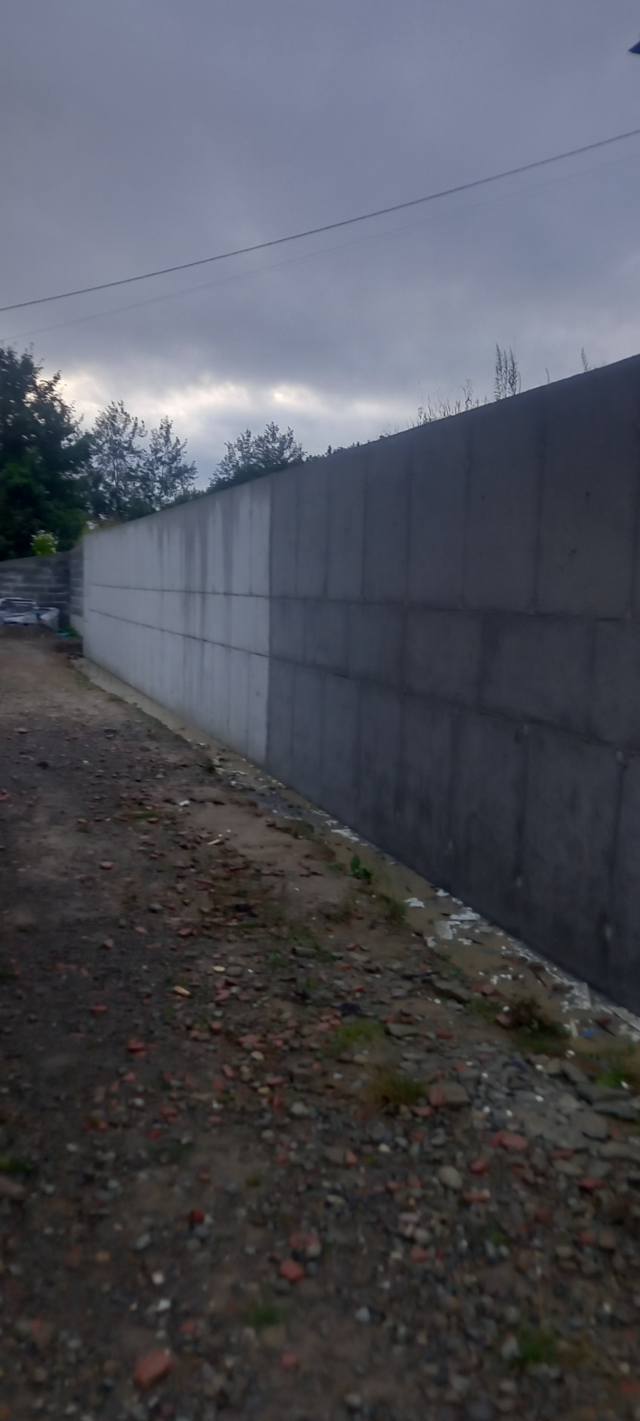 Długi, wysoki, betonowy mur oporowy z widocznymi śladami wilgoci u podstawy, oddzielający teren, z fragmentami gruzu i ziemi wzdłuż jego podstawy, sfotografowany pod pochmurnym niebem.
