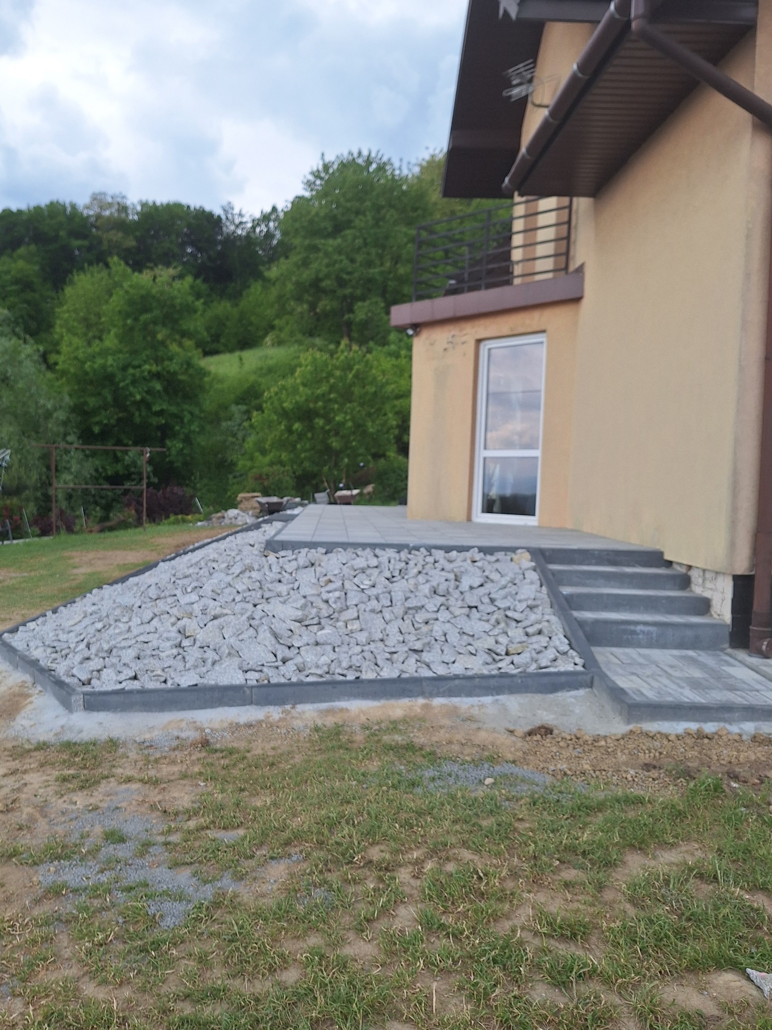 Wykończenie terenu wokół domu z użyciem szarej kostki brukowej i białych kamieni, widoczne schody wejściowe i trawnik.