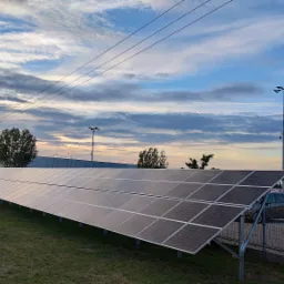 Rząd paneli słonecznych na trawniku, w tle budynek magazynowy i zachmurzone niebo o zmierzchu.