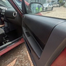 Space Clean - Wnętrze czerwonego samochodu z otwartymi drzwiami, widoczny panel drzwiowy z perforowaną fakturą, fragment deski rozdzielczej i odbicie osoby w lusterku bocznym.