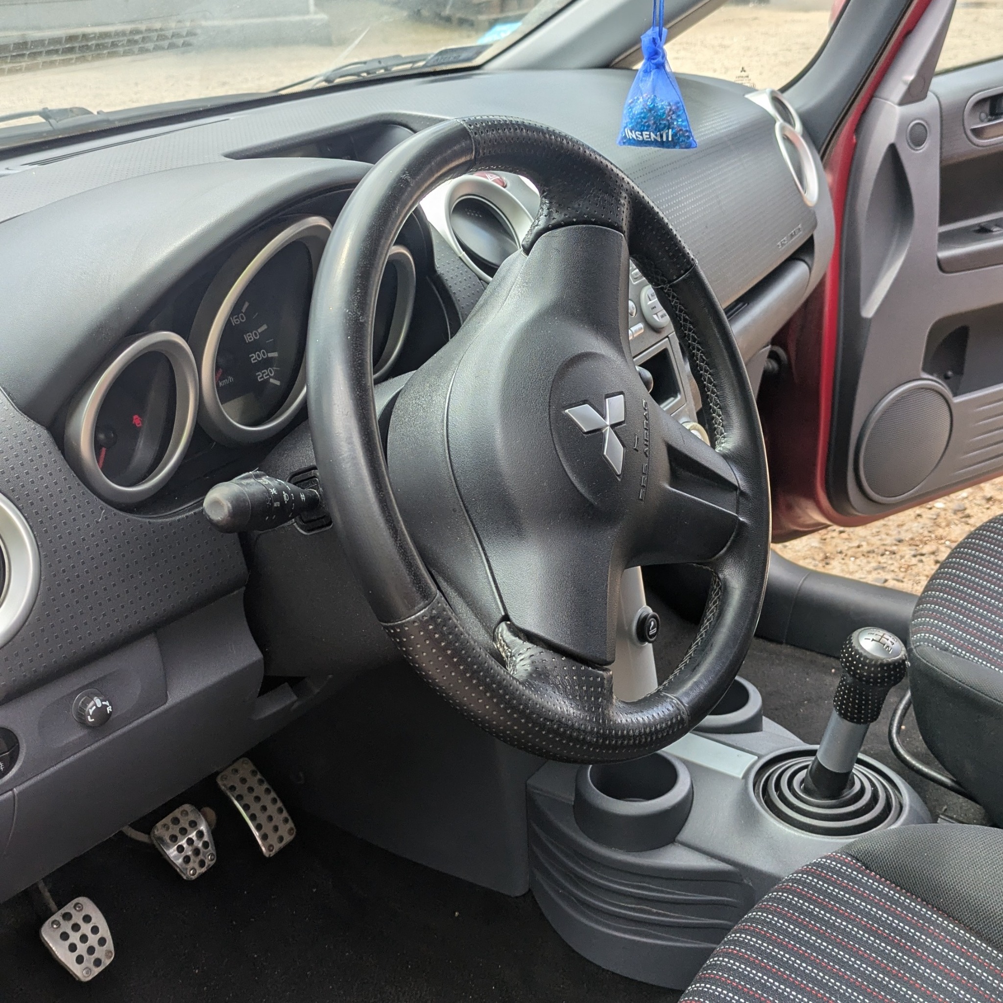 Wnętrze czerwonego samochodu Mitsubishi Colt: kierownica z logo, deska rozdzielcza, pedały z otworami i gałka zmiany biegów z wzorem.