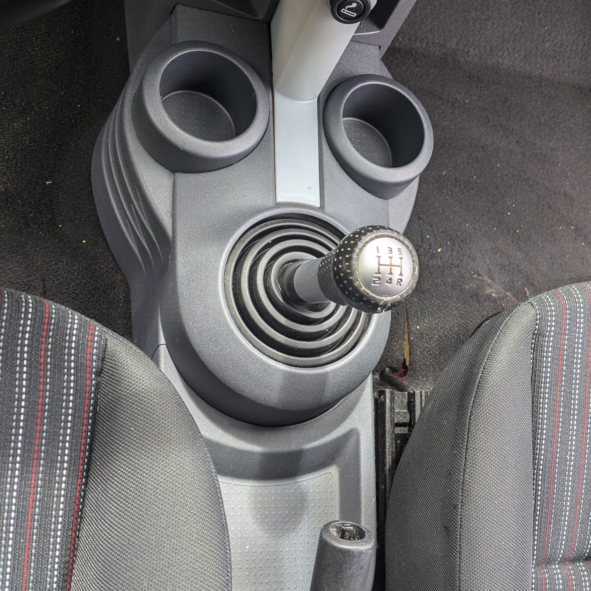 Wnętrze samochodu z widocznym tunelem środkowym, dźwignią zmiany biegów i fragmentami foteli z czarną tapicerką w biało-czerwone paski. Dźwignia zmiany biegów jest lekko zakurzona.