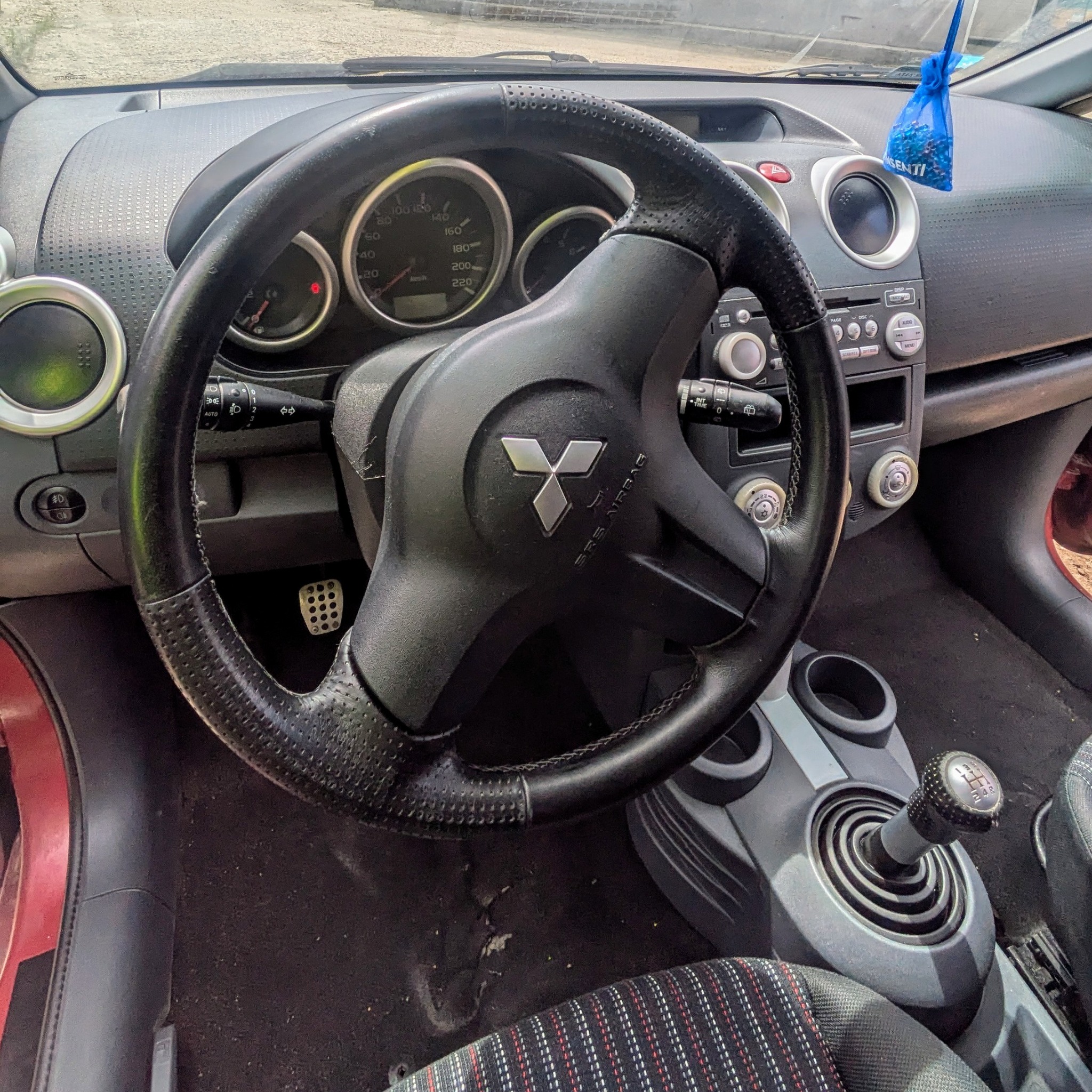 Wnętrze samochodu Mitsubishi Colt: kierownica z logo, deska rozdzielcza z nawiewami i radiem, dźwignia zmiany biegów, widoczny zabrudzony dywanik i odświeżacz powietrza.