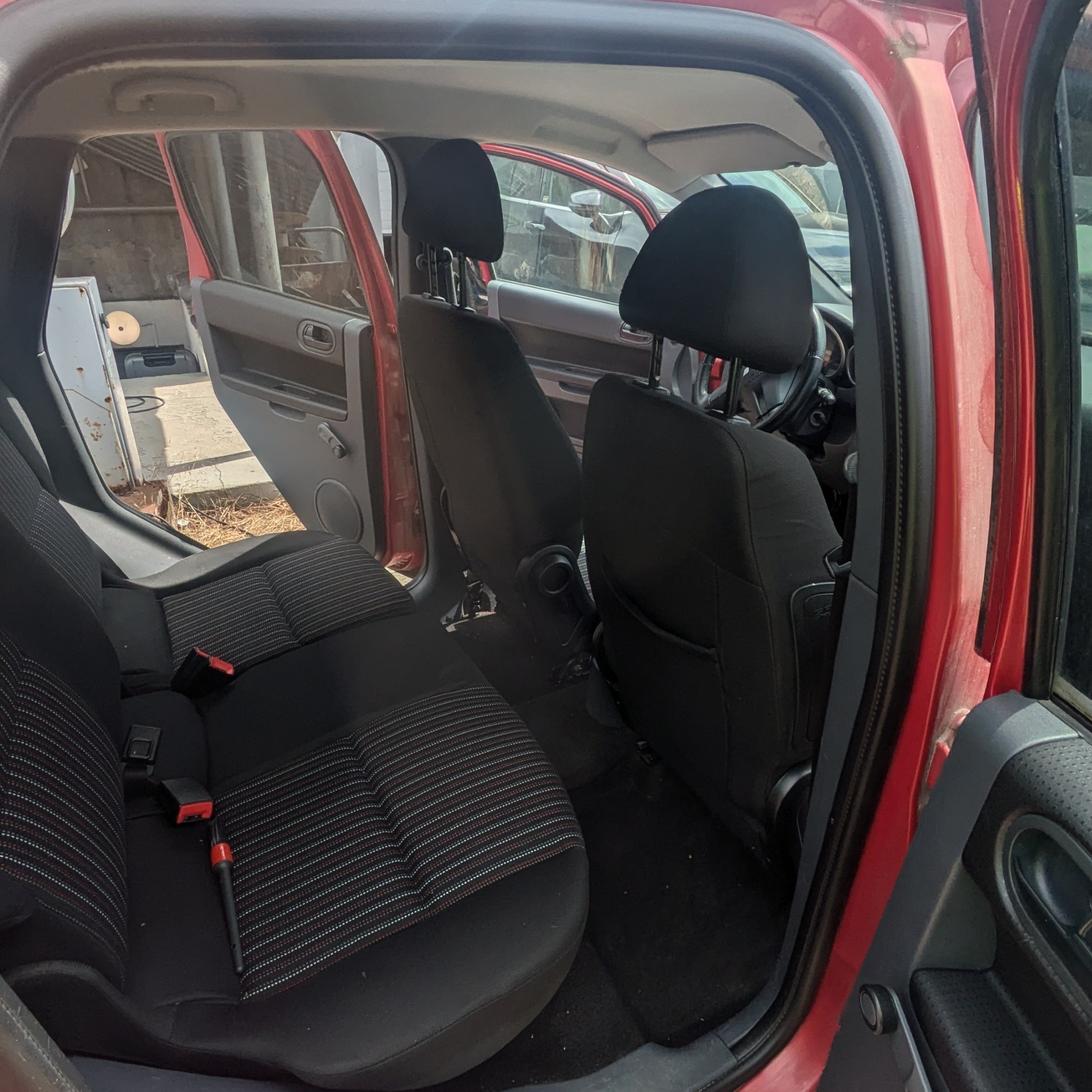 Wnętrze czerwonego samochodu z widocznymi czarnymi fotelami z paskami, na tylnym siedzeniu leży szczotka do czyszczenia detali samochodowych.