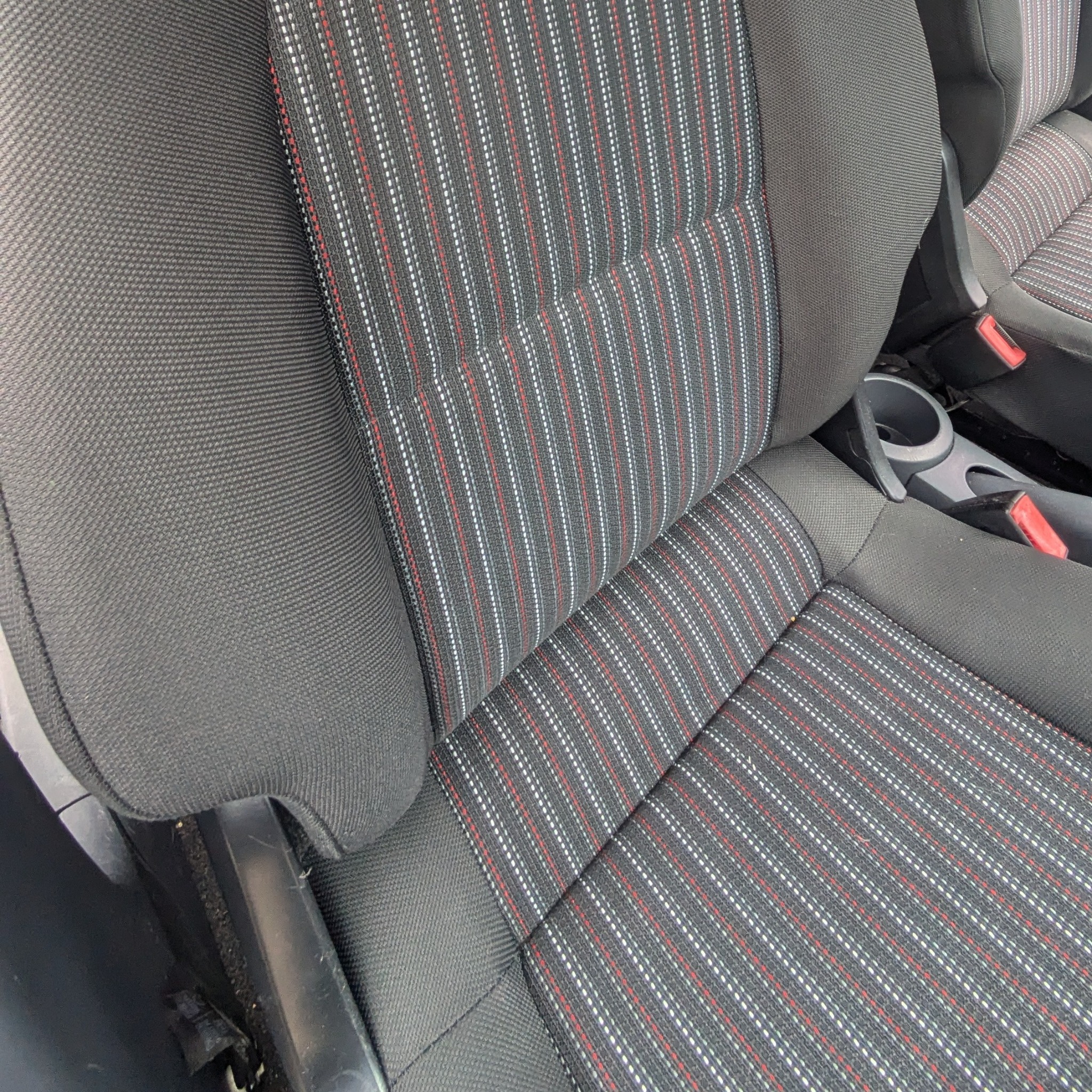 Fragment tapicerowanego fotela samochodowego z czarną, prążkowaną tkaniną w białe i czerwone paski, widoczny uchwyt na napoje i fragment pasów bezpieczeństwa.