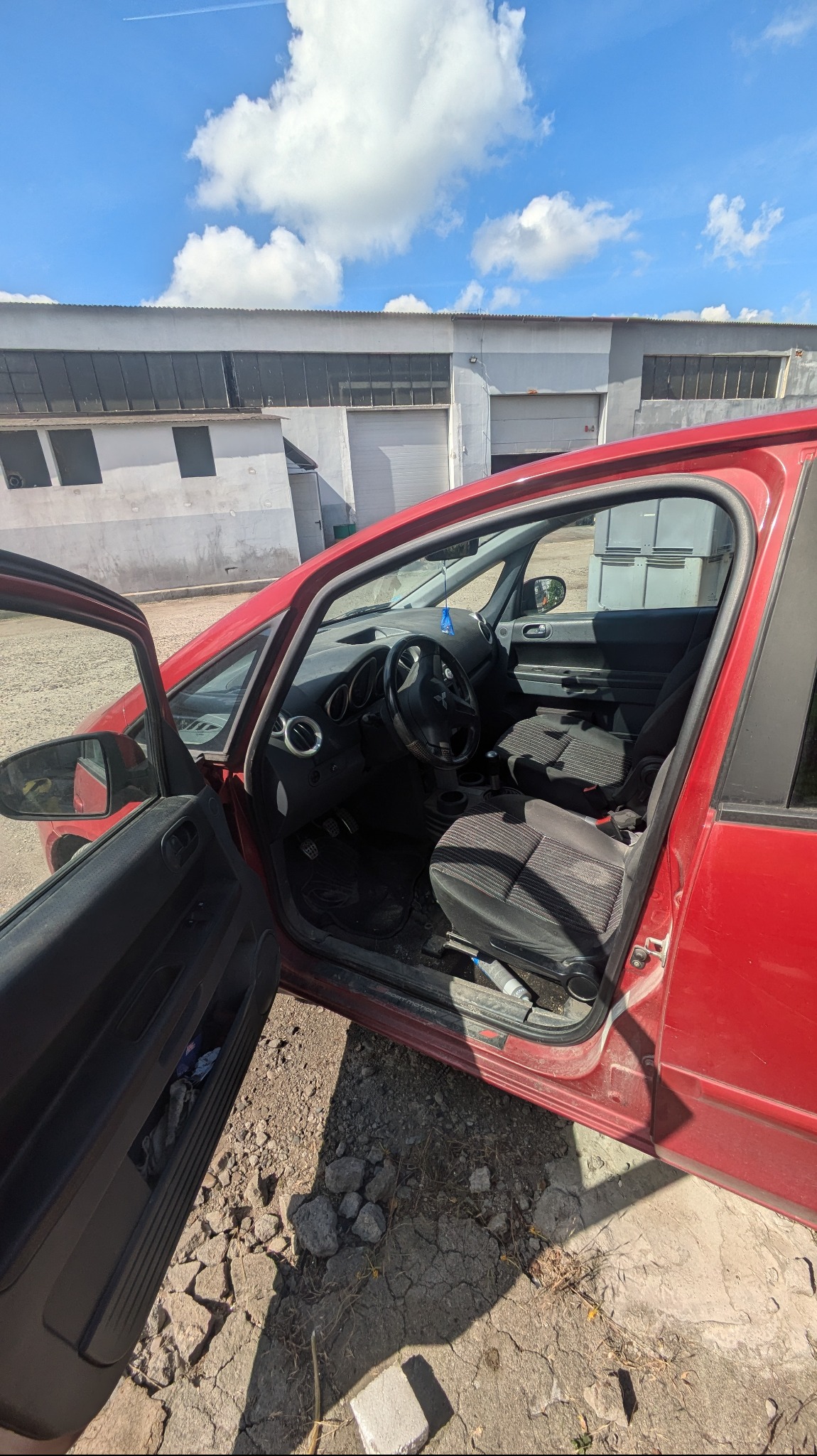 Wnętrze czerwonego samochodu osobowego z otwartymi drzwiami, widoczne czarne fotele z szarą tapicerką w paski, deska rozdzielcza, kierownica, oraz fragment podłoża z gruzem i popękanym asfaltem...