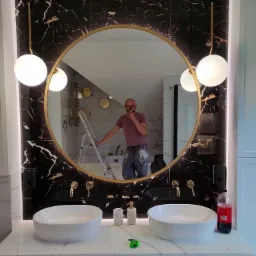 Łazienka w Poznaniu po remoncie: czarne marmurowe płytki, okrągłe lustro z złotą ramą, białe umywalki nablatowe, oświetlenie w stylu modern, widoczny malarz robiący zdjęcie.