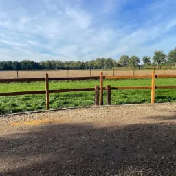 Nowe, drewniane ogrodzenie dwupoziomowe z okrągłymi słupkami, oddzielające trawnik od piaszczystego terenu w Grudziądzu, z widokiem na pole i zadrzewienia w tle.