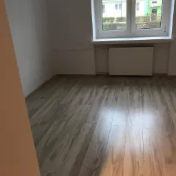 Jasne panele podłogowe w pokoju z białymi ścianami i oknem z widokiem na budynek mieszkalny.