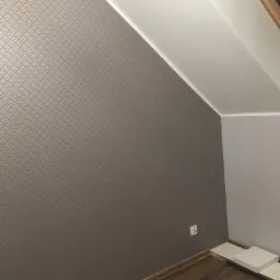 Ściana w pokoju na poddaszu pokryta tapetą w geometryczny wzór, widoczny fragment drewnianej podłogi i białe listwy przypodłogowe.