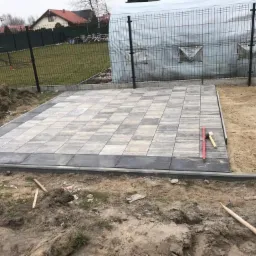 Układanie kostki brukowej na tarasie, widoczne narzędzia: poziomica i gumowy młotek, obrzeża tarasu wykończone ciemniejszą kostką, w tle siatka ogrodzeniowa i budynek.