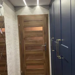 Nowoczesny przedpokój z drewnianymi drzwiami z przeszkleniami i granatową szafą z złotymi uchwytami, oświetlenie sufitowe LED.