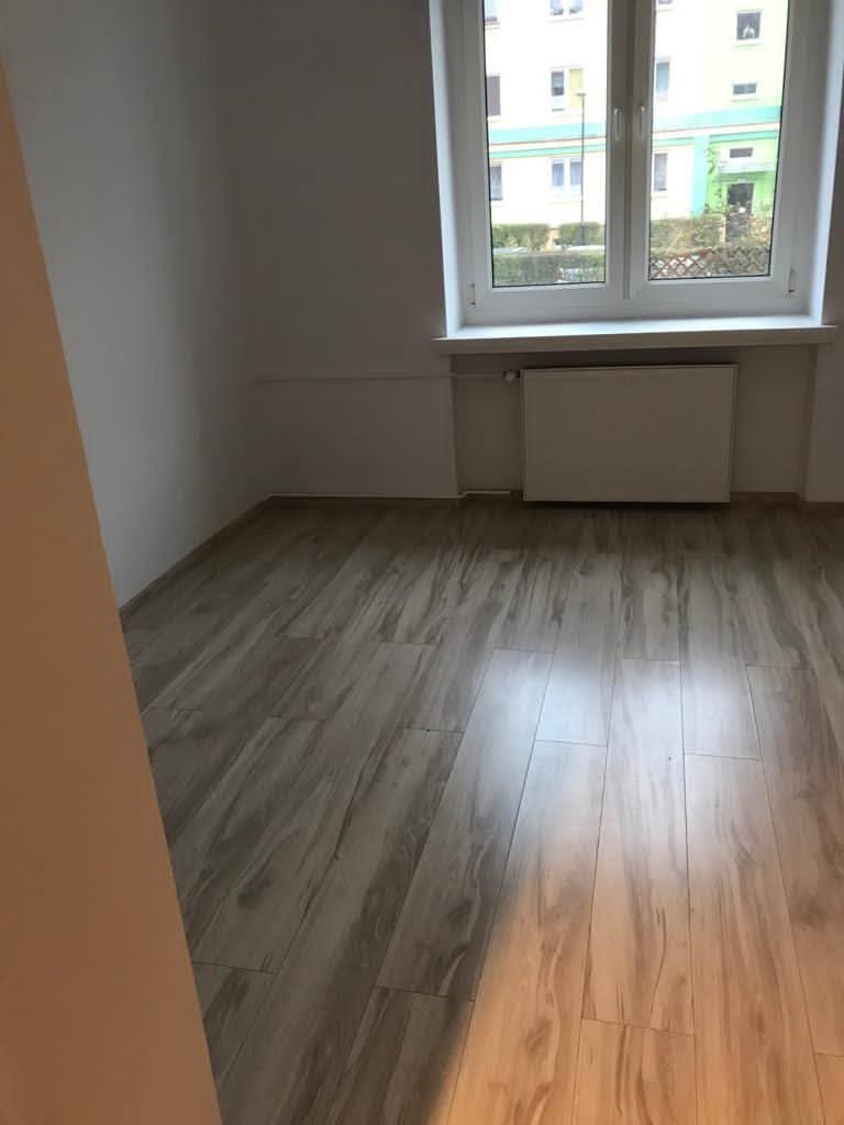 Pokój po remoncie z nową podłogą z paneli imitujących drewno, widok na okno z białymi ramami i grzejnik pod nim.