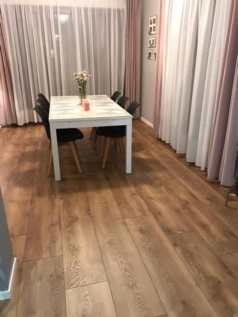 Jasna, drewniana podłoga z paneli, widoczny stół z czarnymi krzesłami, wazon z kwiatami i zasłony w tle.