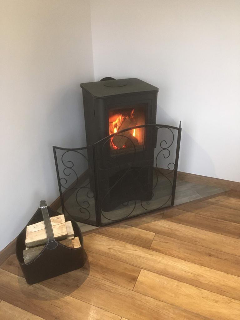 Kompaktowy piec wolnostojący z palącym się drewnem, zabezpieczony ozdobnym, metalowym parawanem; obok skórzany kosz z przygotowanym drewnem opałowym, na panelach podłogowych w odcieniach brązu.