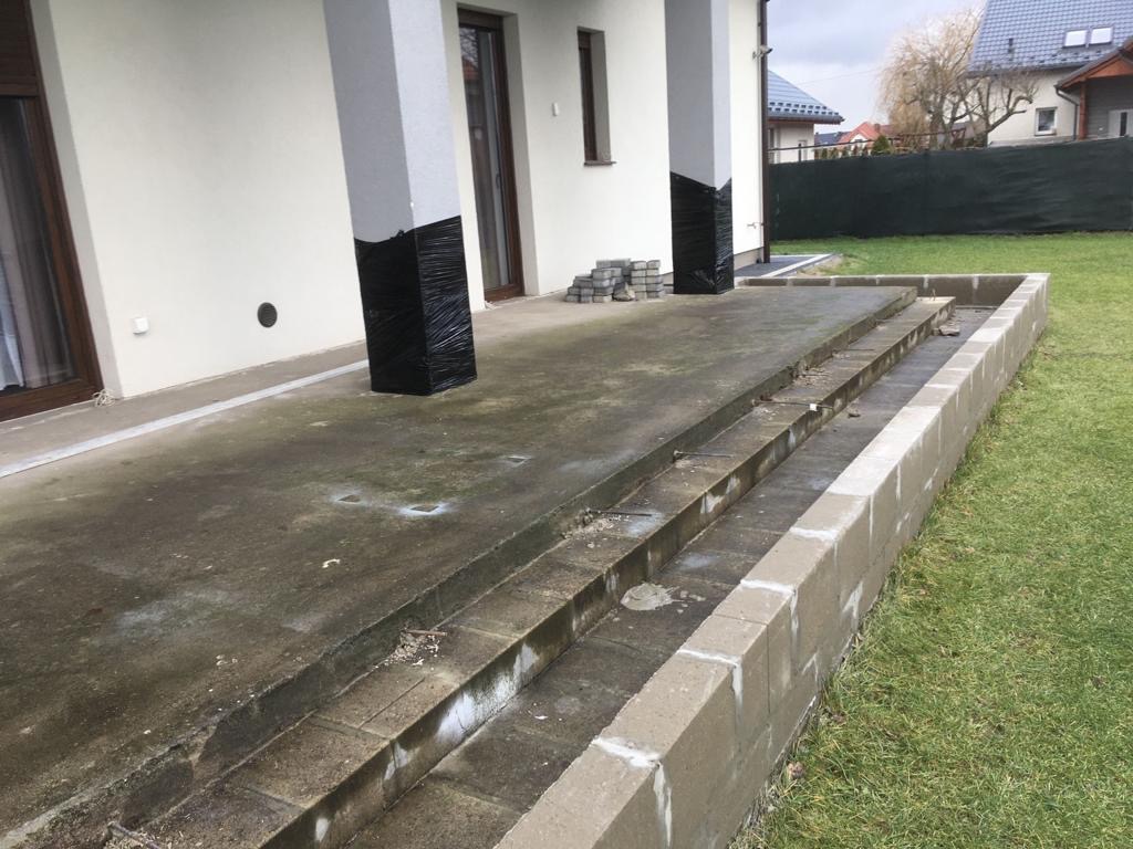 Budowa betonowego tarasu z trzema schodami i zabezpieczonymi folią kolumnami przy białym domu z brązowymi oknami.