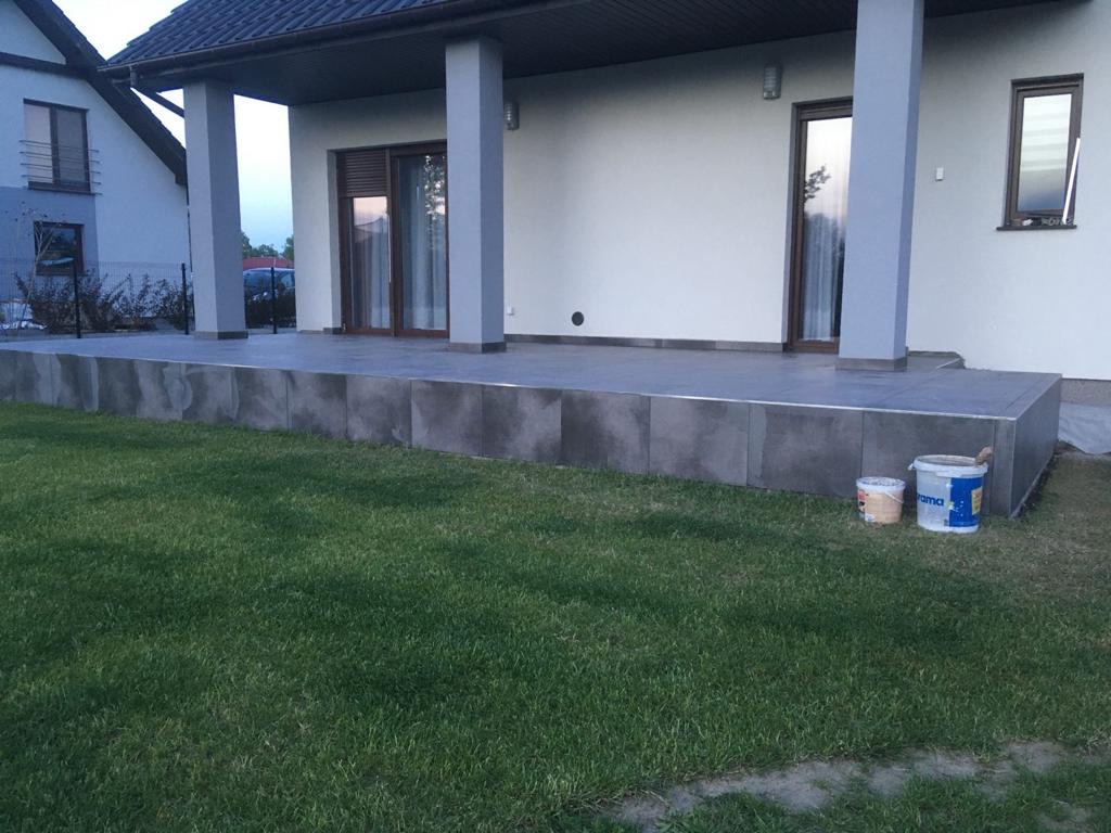 Wykończony taras z szarych płytek ceramicznych, widoczne dwa wiadra po farbie, trawnik w pierwszym planie.