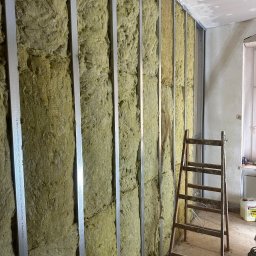 Firma Handlowo-Usługowa Artur Adamczyk - Ocieplenie ściany wełną mineralną, widoczne metalowe profile i drabina. Etap prac przed zakryciem izolacji płytami gipsowo-kartonowymi. Surowy stan wykończenia.