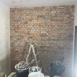 Firma Handlowo-Usługowa Artur Adamczyk - Prace wykończeniowe: mur z cegły, drabina malarska, wiadra z farbą i narzędzia na stole. Mężczyzna w trakcie pracy nad ścianą z cegły.