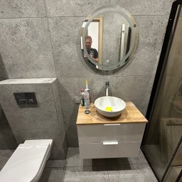 Firma Handlowo-Usługowa Artur Adamczyk - Nowoczesna łazienka z szarą płytką, okrągłym lustrem z podświetleniem LED, wiszącą szafką z umywalką i toaletą. Widoczne detale wykończenia.