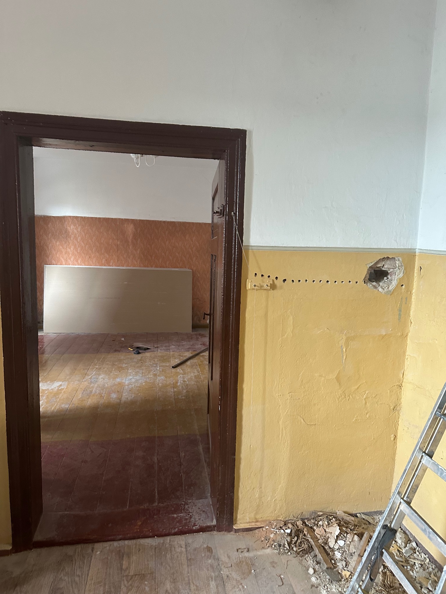 Pokój w trakcie remontu: żółta ściana z dziurą i śladami po listwie, widoczne płyty karton-gips w przejściu, drewniana podłoga. Fragment drabiny po prawej.