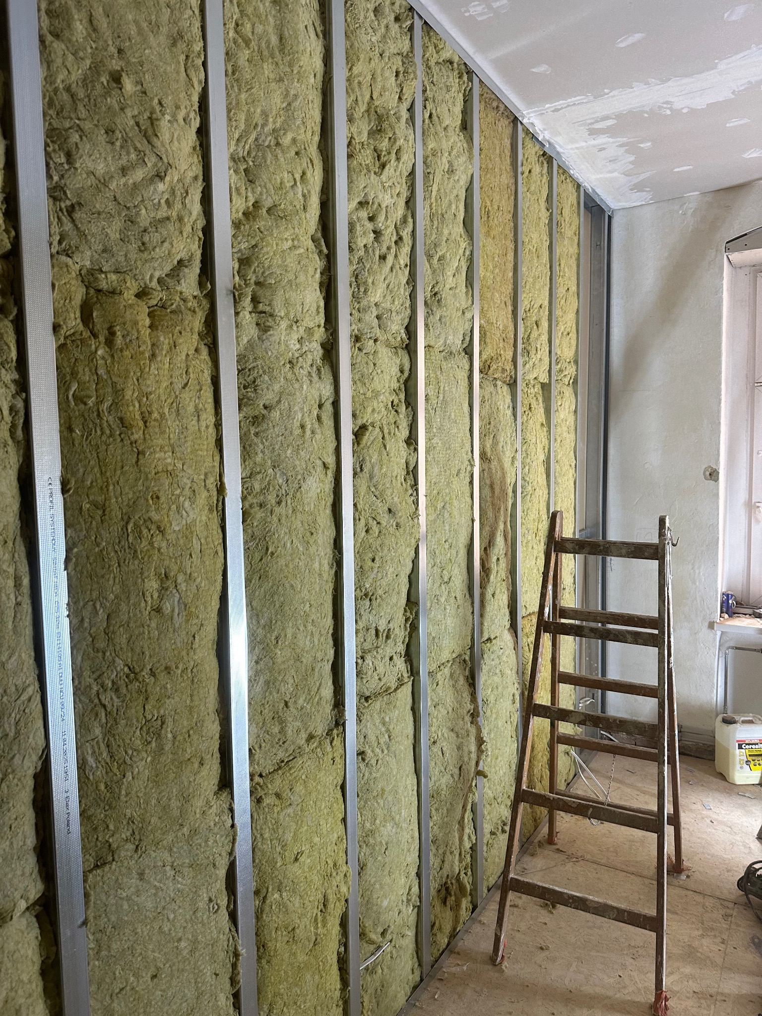 Ocieplenie ściany wełną mineralną, widoczne metalowe profile i drabina. Etap prac przed zakryciem izolacji płytami gipsowo-kartonowymi. Surowy stan wykończenia.