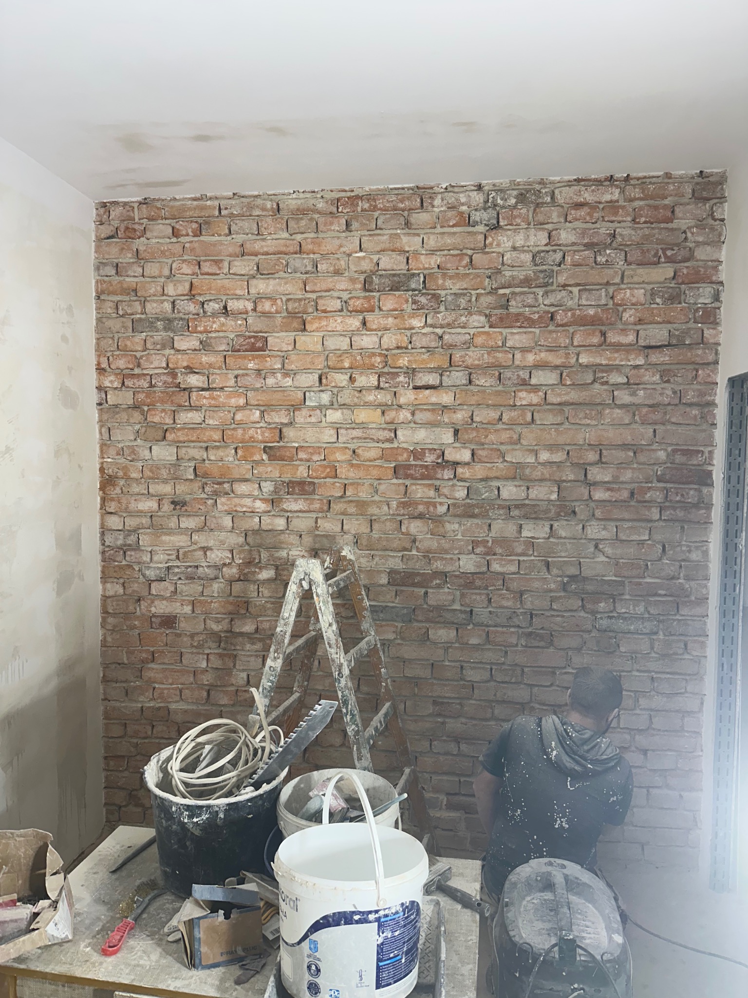 Prace wykończeniowe: mur z cegły, drabina malarska, wiadra z farbą i narzędzia na stole. Mężczyzna w trakcie pracy nad ścianą z cegły.
