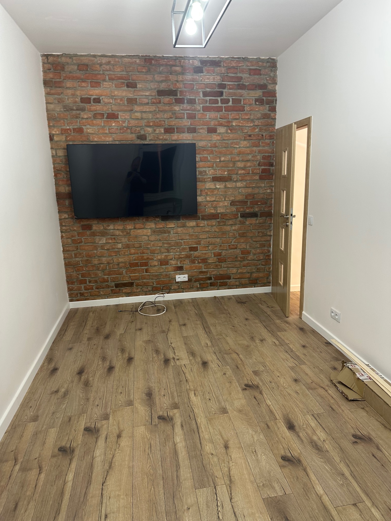 Pokój po remoncie: drewniana podłoga, ściana z cegły z telewizorem, białe ściany. Widoczne listwy przypodłogowe i otwarte drzwi. Nowoczesne oświetlenie sufitowe.