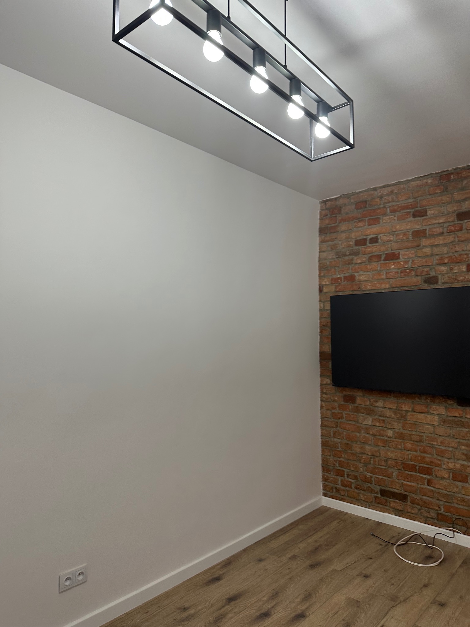 Minimalistyczne wnętrze: gładka, biała ściana kontrastuje z ceglaną ścianą z telewizorem. Parkiet i nowoczesna lampa sufitowa dopełniają wystrój.