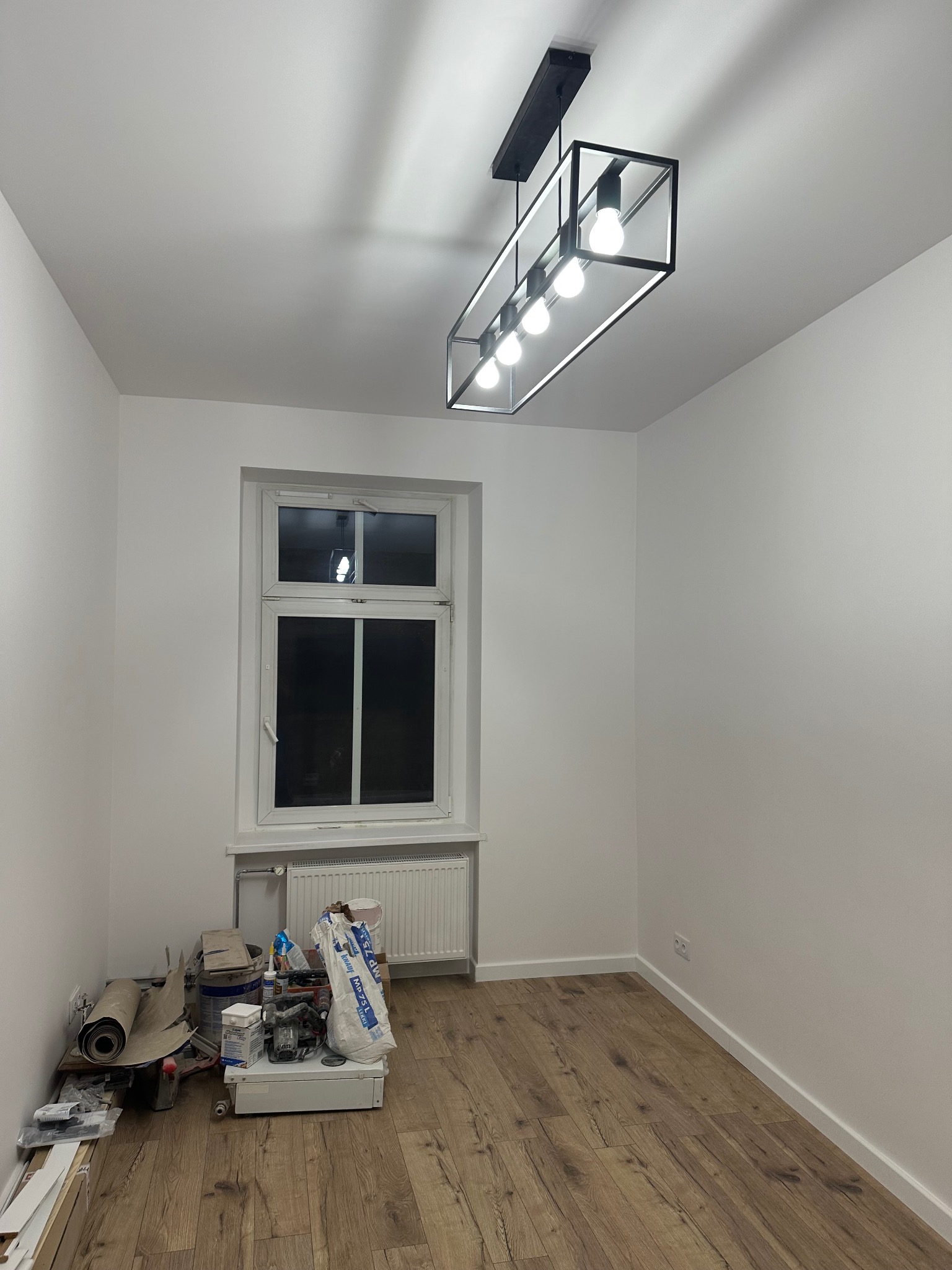 Pokój po remoncie: świeżo pomalowane białe ściany, ułożone panele podłogowe w odcieniach brązu. Widoczne narzędzia i materiały malarskie przy ścianie. Nowoczesna lampa sufitowa.