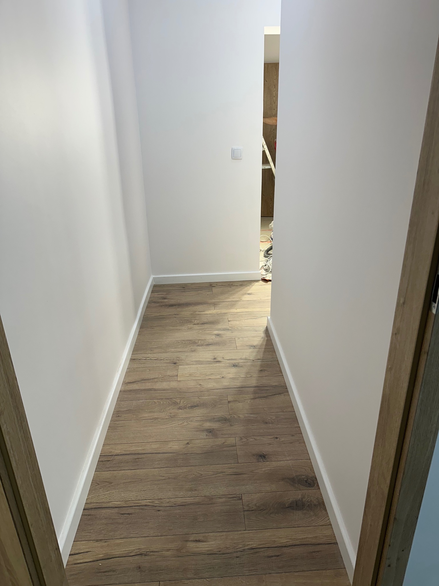 Wąski korytarz z panelami podłogowymi w odcieniach brązu i świeżo pomalowanymi na biało ścianami. Widoczny fragment ościeżnicy drzwi i biała listwa przypodłogowa.