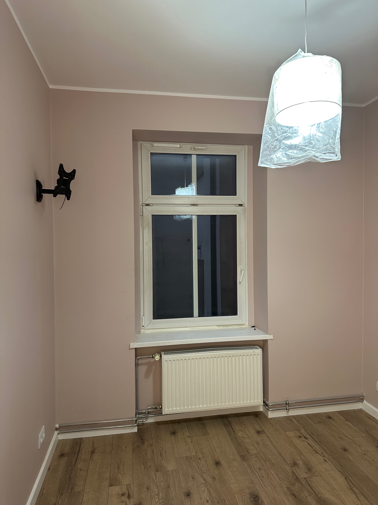 Świeżo pomalowane ściany w odcieniach różu w pokoju z drewnianą podłogą, białe okno i grzejnik. Na ścianie uchwyt na telewizor, lampa w folii ochronnej.