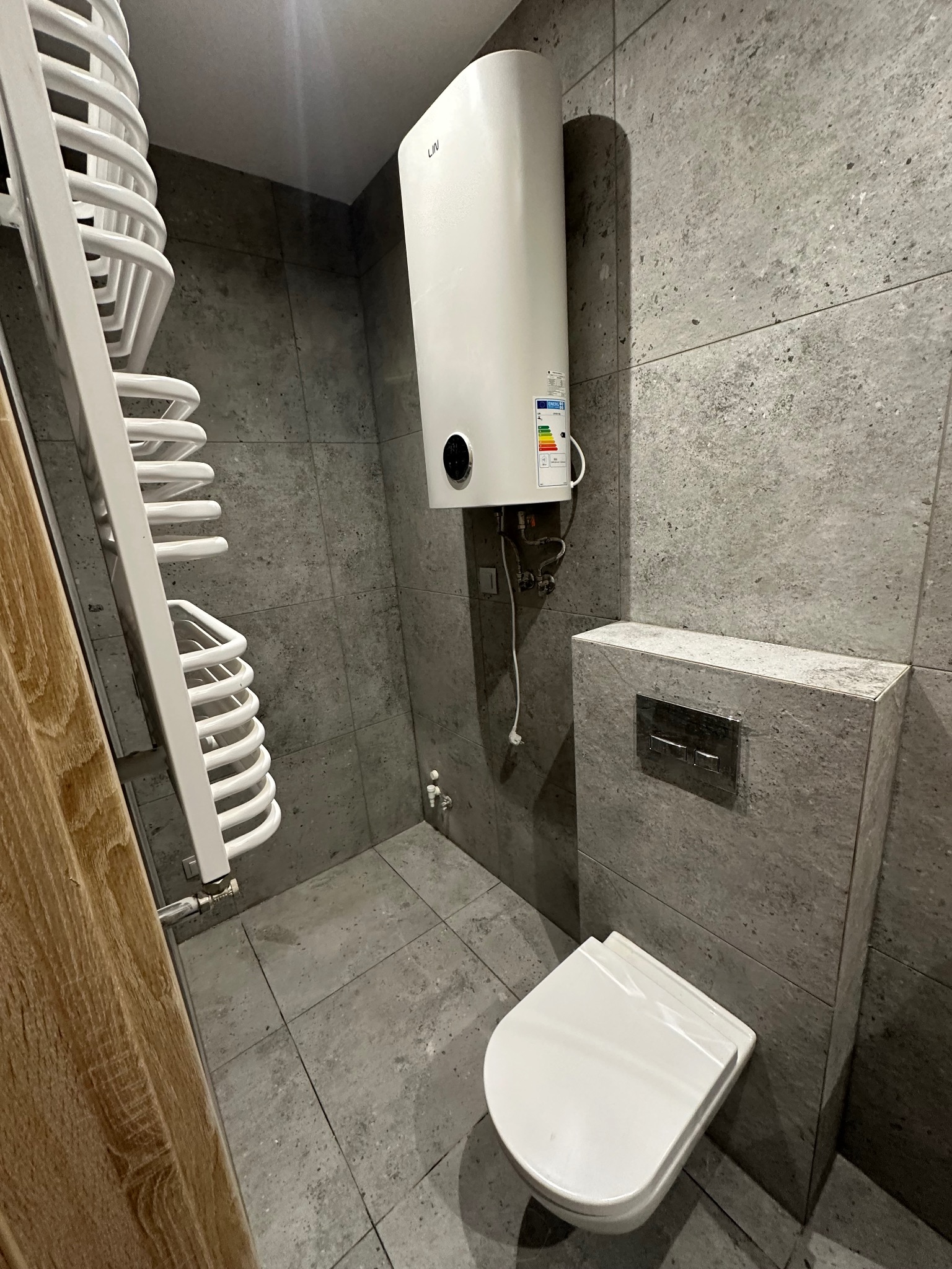 Nowoczesna łazienka z szarymi płytkami, podwieszaną toaletą i białym grzejnikiem drabinkowym. Widoczny bojler elektryczny na ścianie.