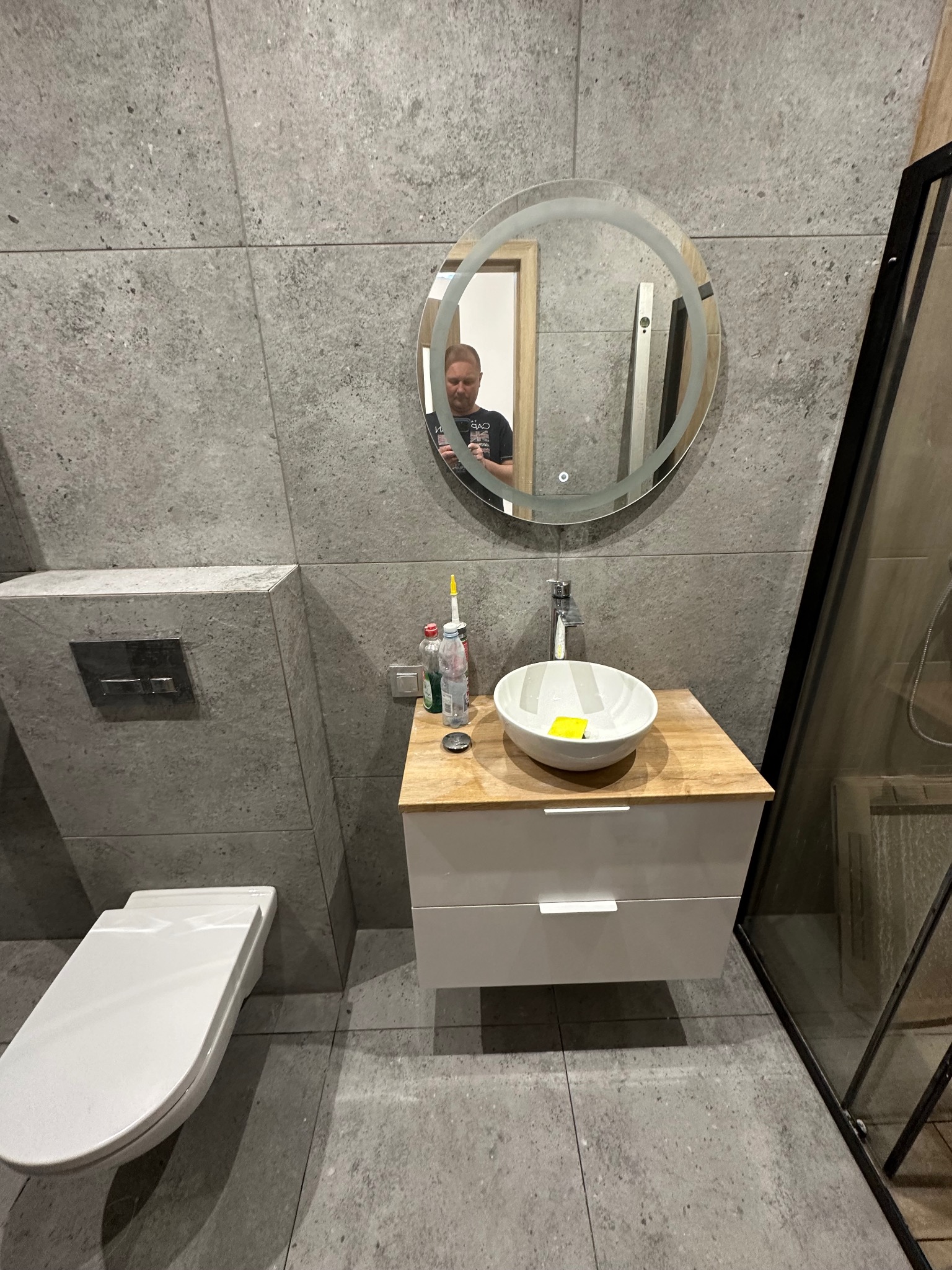 Nowoczesna łazienka z szarą płytką, okrągłym lustrem z podświetleniem LED, wiszącą szafką z umywalką i toaletą. Widoczne detale wykończenia.