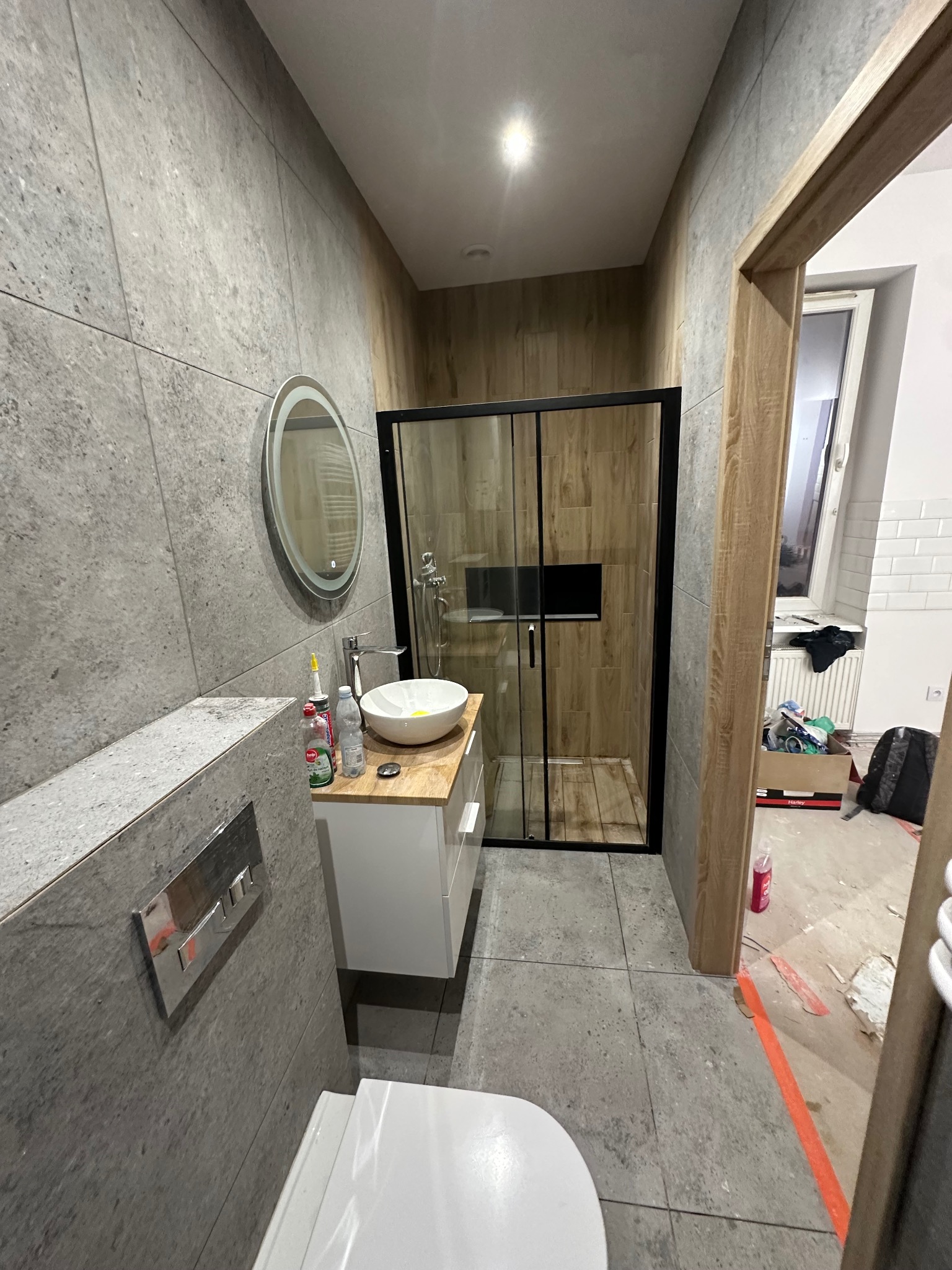 Wykończona łazienka z szarą płytką, okrągłym lustrem z podświetleniem LED, kabiną prysznicową w czarnej ramie i elementami białej ceramiki. Widoczne ślady po remoncie.