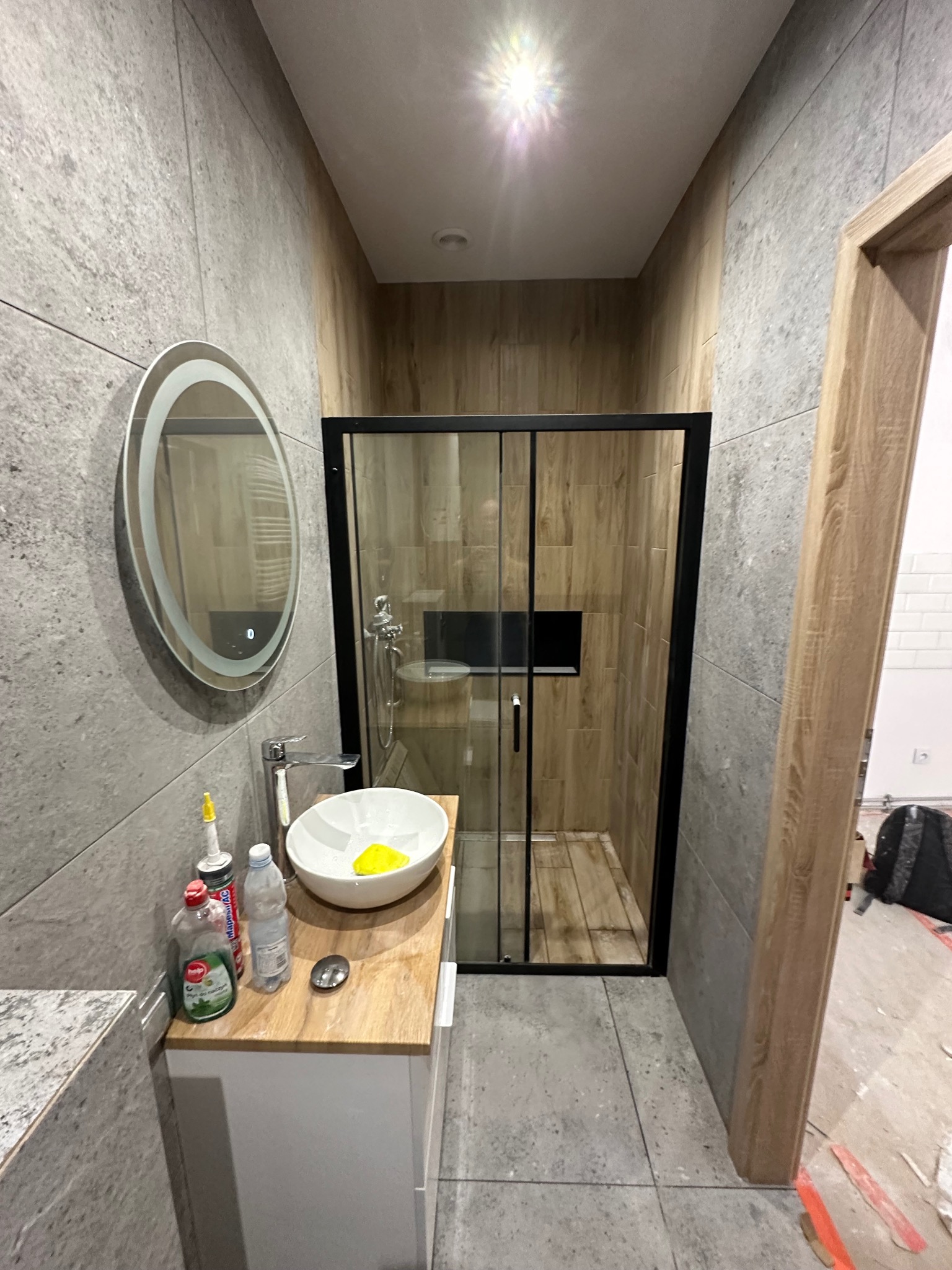 Wykończona łazienka z kabiną prysznicową w stylu drewna i szarą płytką. Okrągłe lustro z podświetleniem nad umywalką. Widoczne detale wykończeniowe.