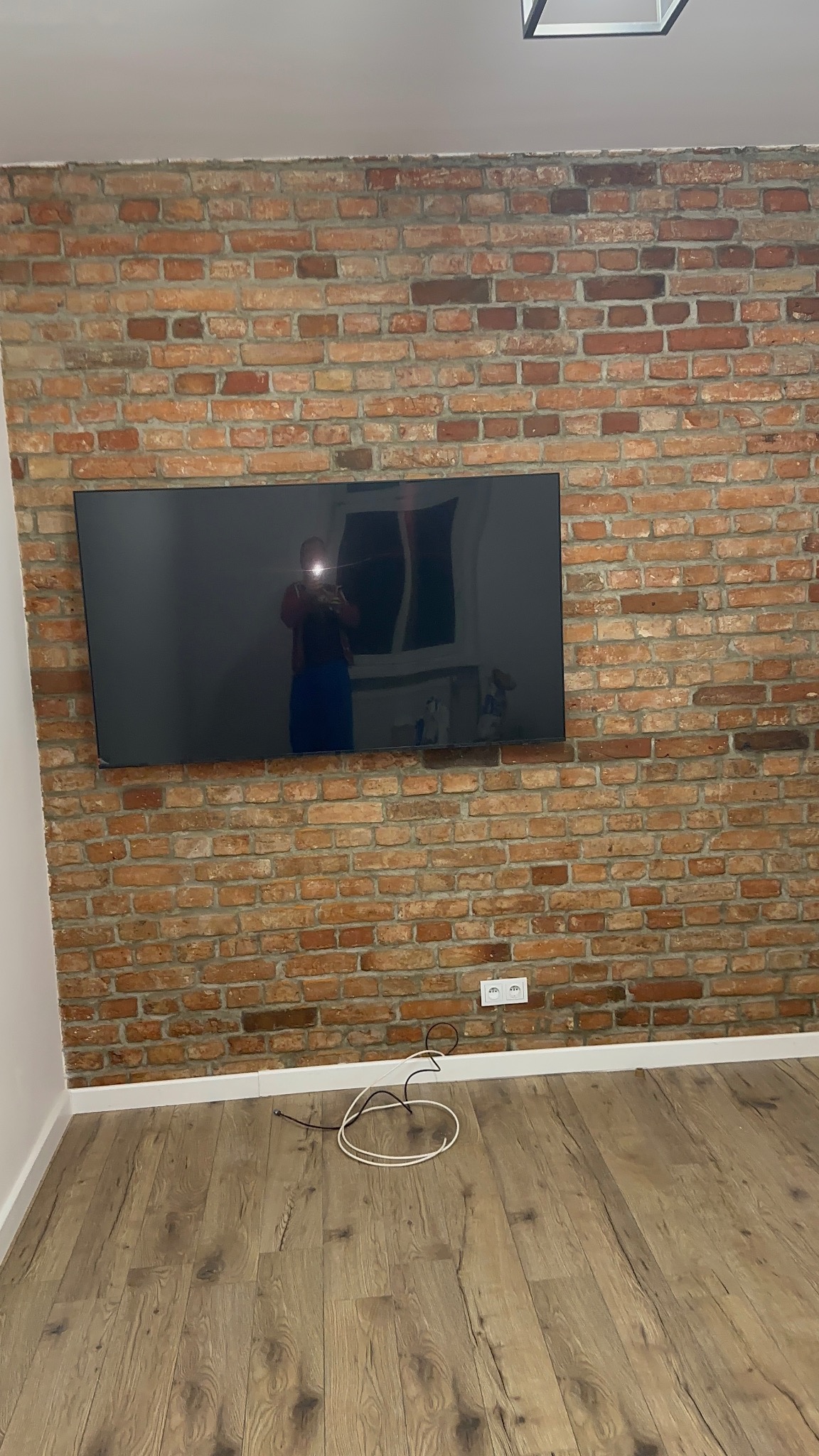Telewizor zawieszony na ścianie z czerwonej cegły, widoczne gniazdka elektryczne i podłoga z paneli. W odbiciu ekranu widoczna osoba robiąca zdjęcie.