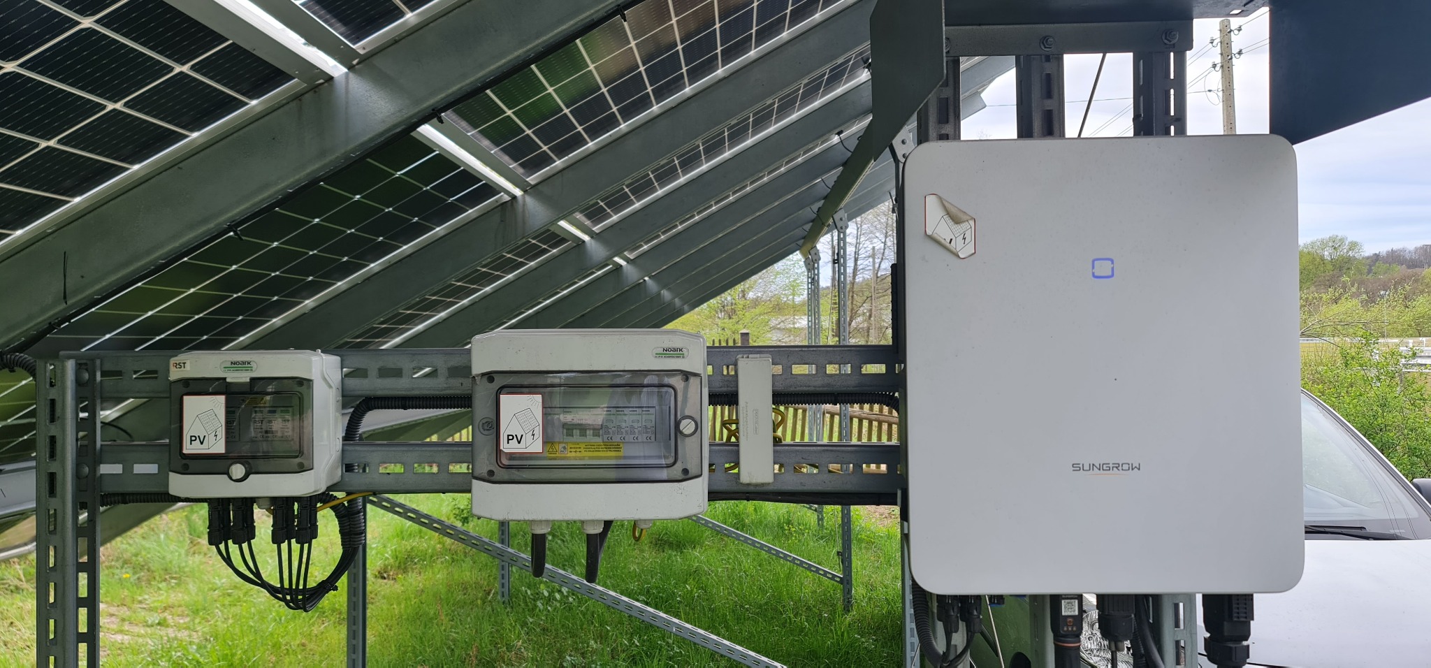 Instalacja fotowoltaiczna: panele, falownik Sungrow i zabezpieczenia Noark zamontowane na konstrukcji wsporczej, widok z poziomu gruntu.