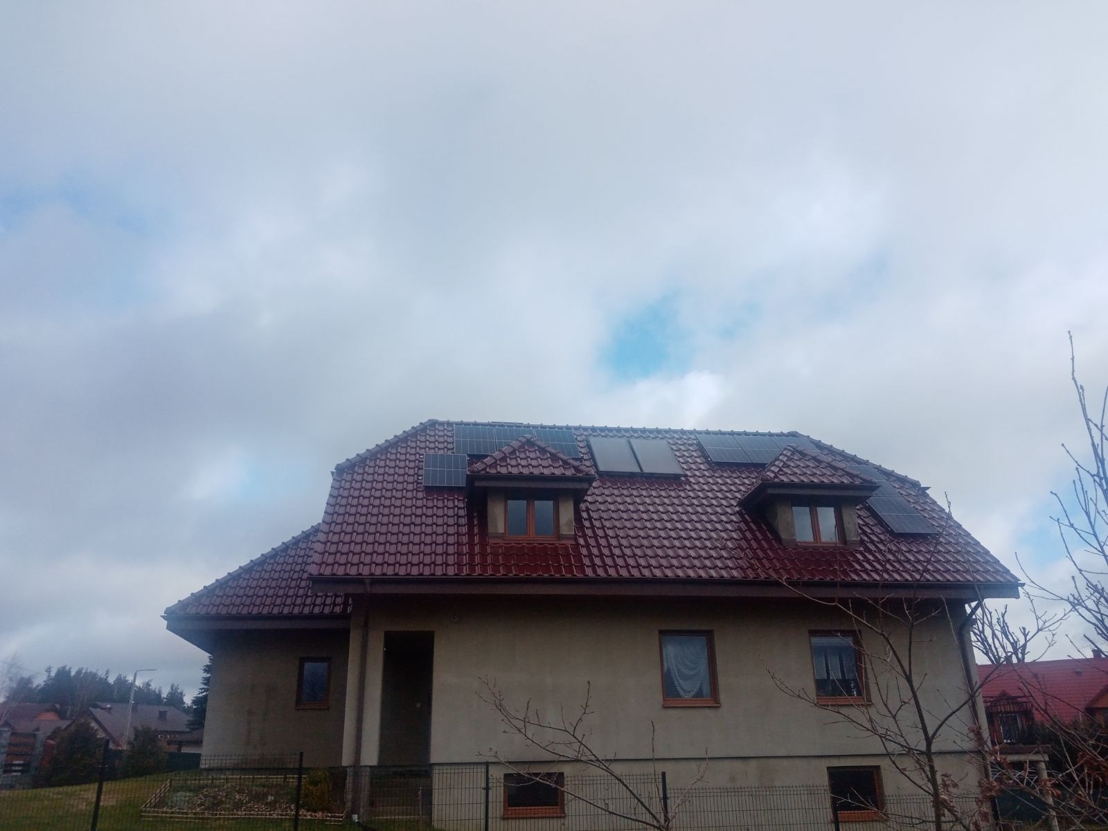 Dom jednorodzinny z ciemnoczerwoną dachówką i panelami słonecznymi na dachu oraz nad oknami dachowymi, widok z dołu w pochmurny dzień.