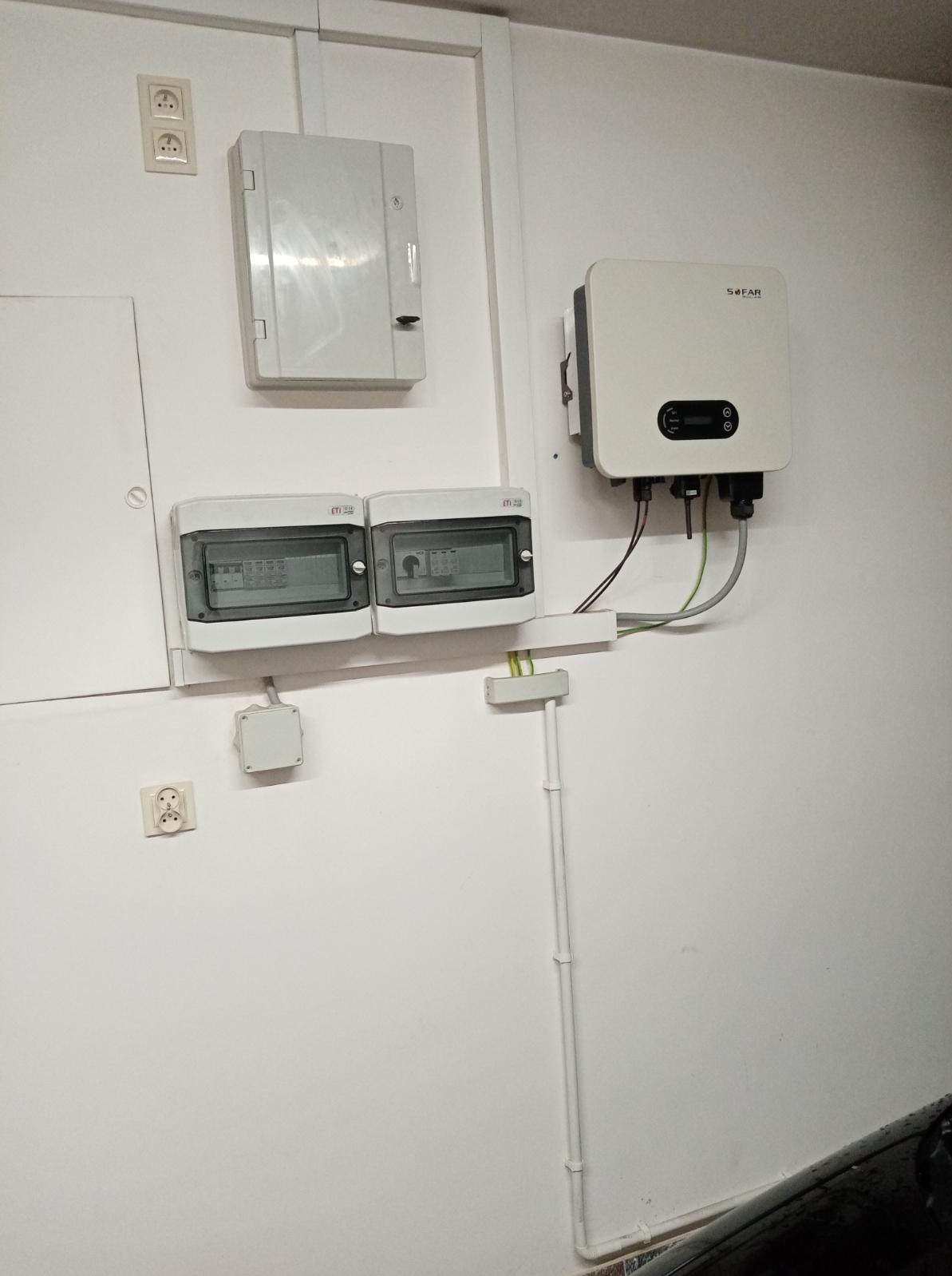 Biały inwerter marki SOFAR zamontowany na ścianie, poniżej dwie szare skrzynki bezpiecznikowe, obok białe puszki elektryczne i rury z kablami.