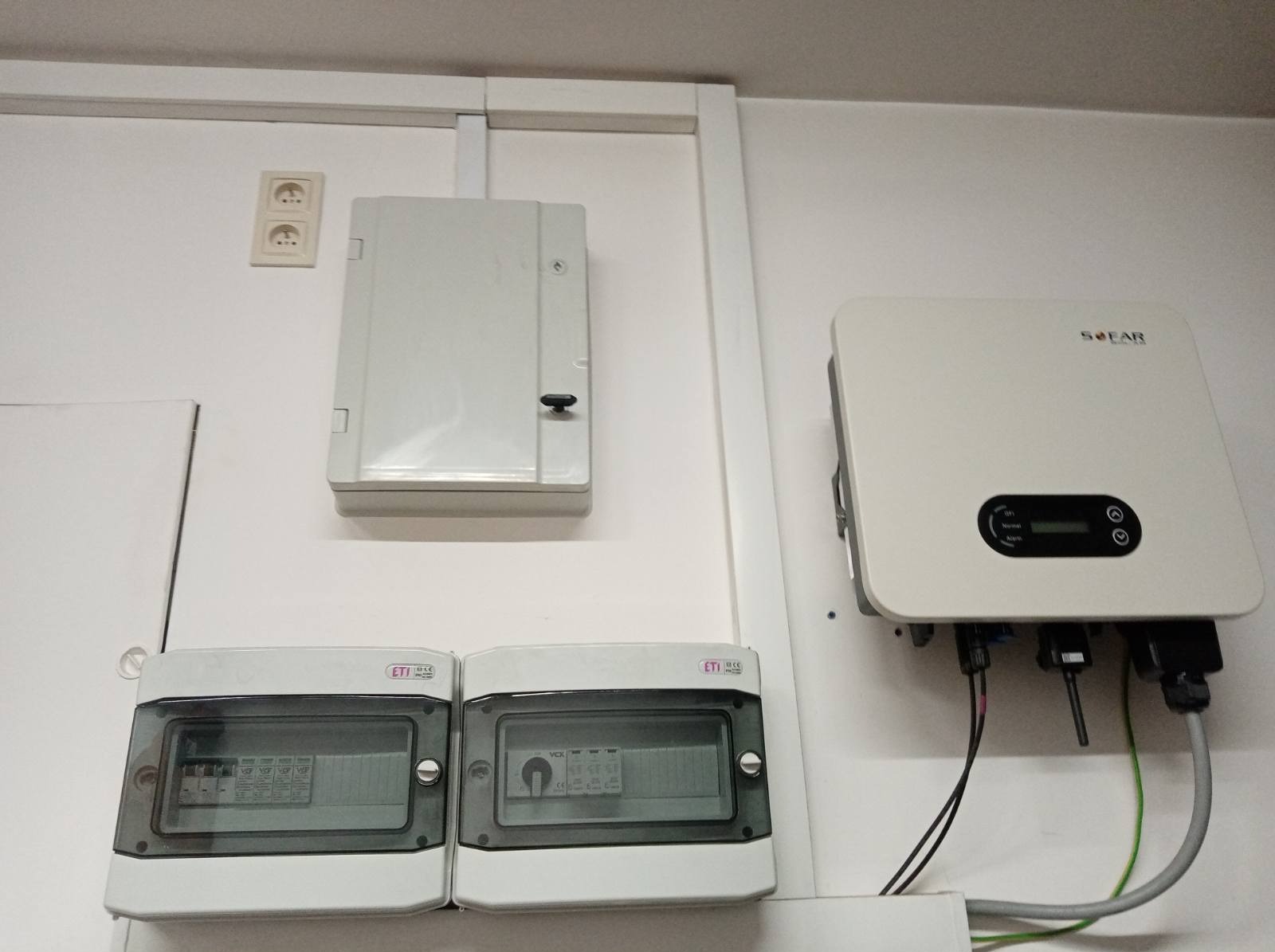 Biała ściana z zamontowanym inwerterem solarnym marki SOFAR, dwoma bezpiecznikami ETI i skrzynką elektryczną. Widoczne kable i przewody. Gniazdko elektryczne nad skrzynką.