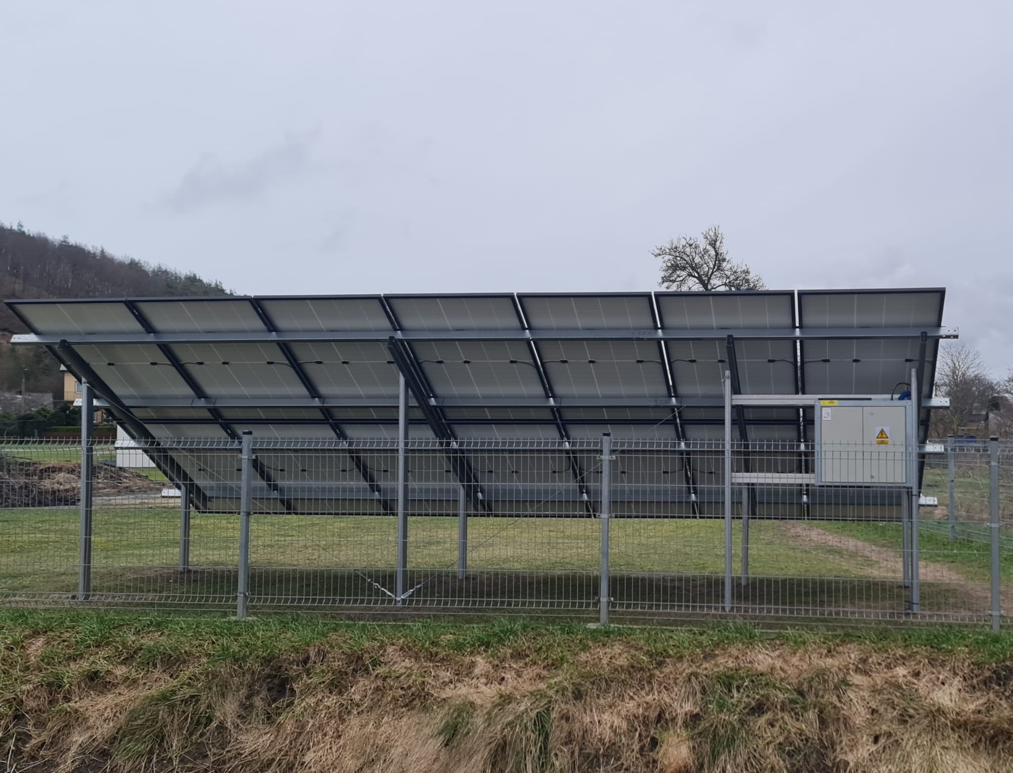 Widok od tyłu na panele słoneczne zamontowane na metalowej konstrukcji, ogrodzone siatką, z szarą skrzynką elektryczną z symbolem ostrzegawczym widoczną po prawej stronie, na tle pochmurnego nieba...