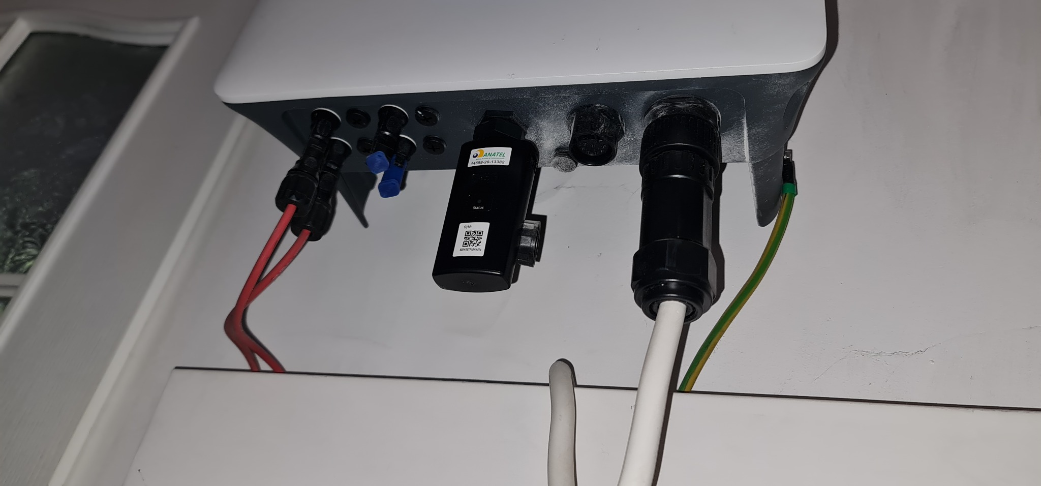 Przyłącza elektryczne do falownika instalacji fotowoltaicznej, widoczne kable, wtyczki, moduł komunikacji z QR kodem oraz przewód uziemiający.