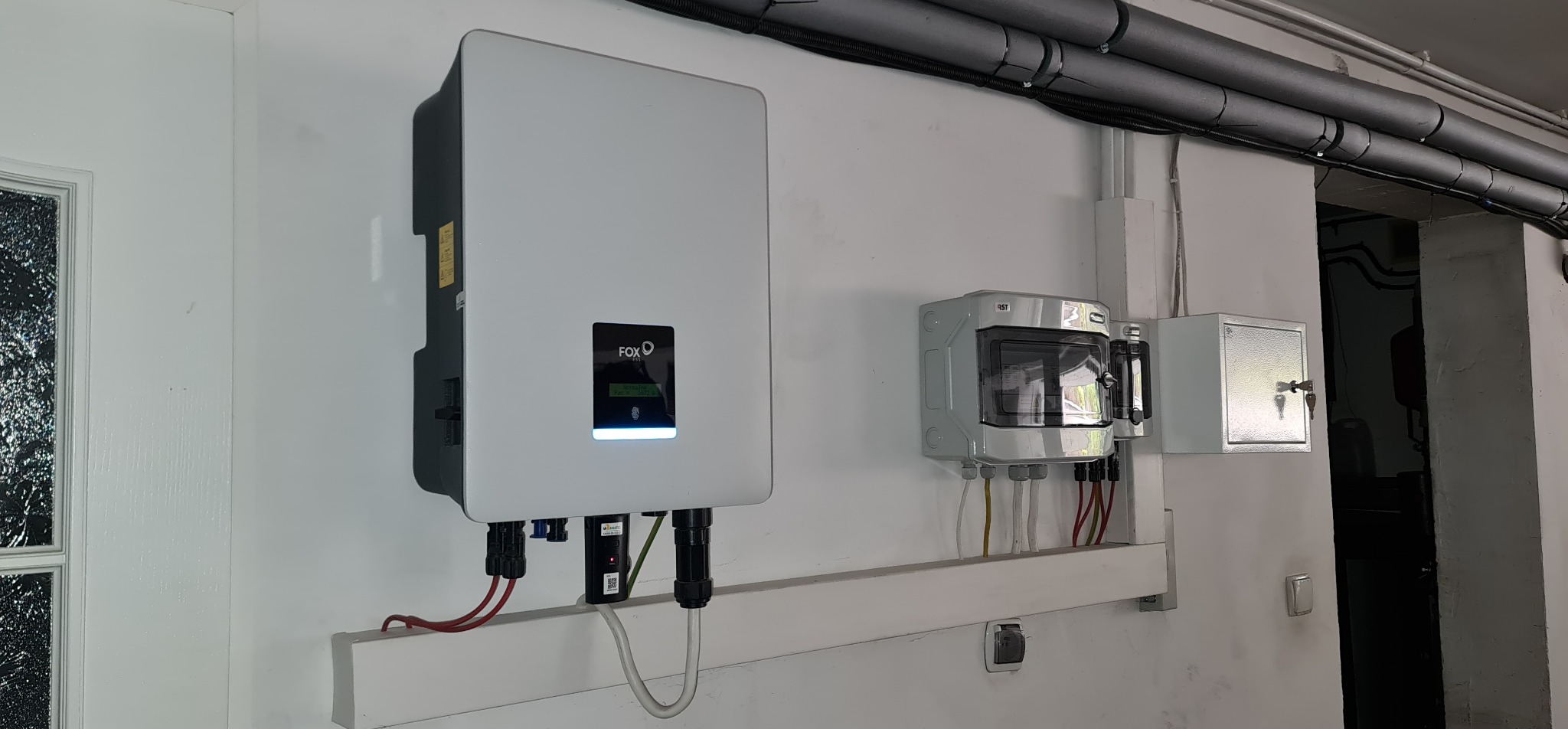 Biały inwerter firmy FoxESS zamontowany na ścianie obok skrzynki bezpiecznikowej i małej metalowej szafki z kluczem, widoczne kable i rury instalacyjne