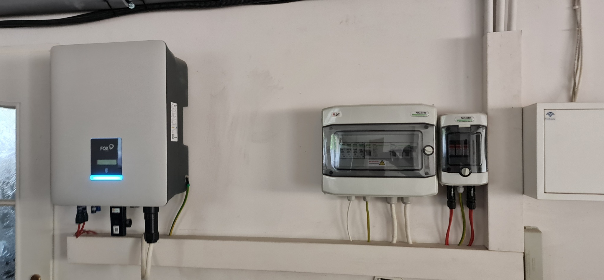 Zamontowany inwerter fotowoltaiczny FoxESS z podłączonymi przewodami, obok skrzynka bezpiecznikowa Noark RST i ogranicznik przepięć Noark, na białej ścianie.