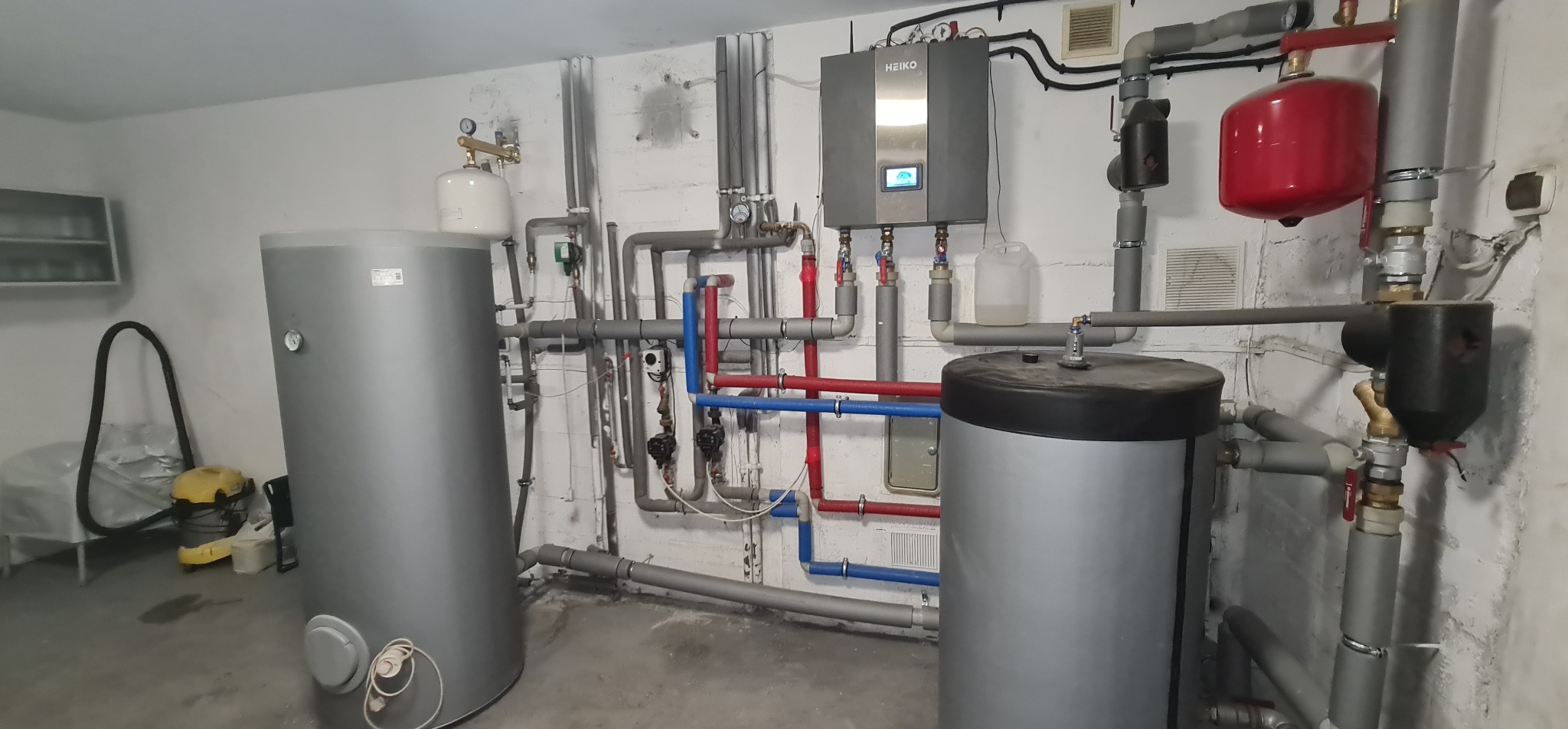 Instalacja centralnego ogrzewania z dwoma szarymi zbiornikami, kotłem marki Heiko i rozbudowaną siecią rur w kolorach szarym, czerwonym i niebieskim, znajdująca się w piwnicy z widocznym...