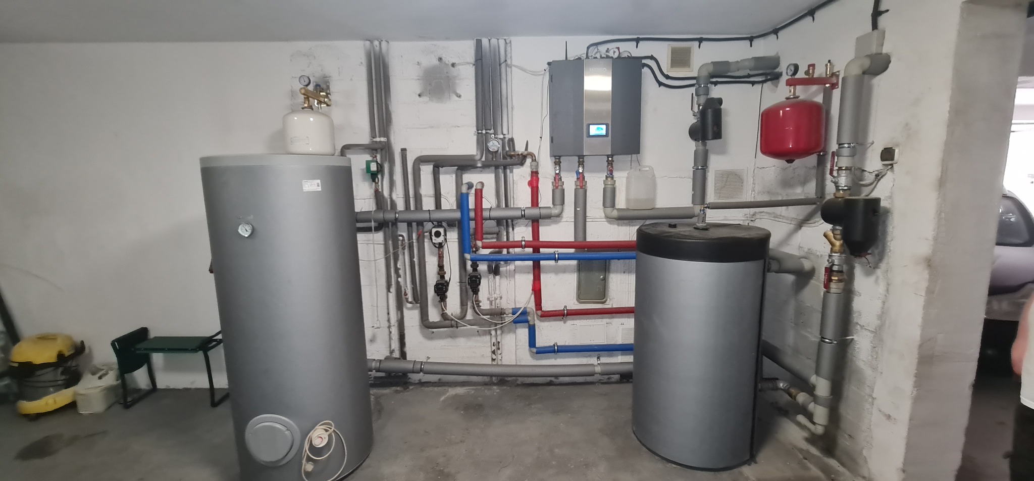 Instalacja grzewcza w piwnicy: dwa szare zbiorniki z podłączeniami rur w kolorze szarym, czerwonym i niebieskim, regulator na ścianie, widoczny żółty odkurzacz i krzesło w tle.