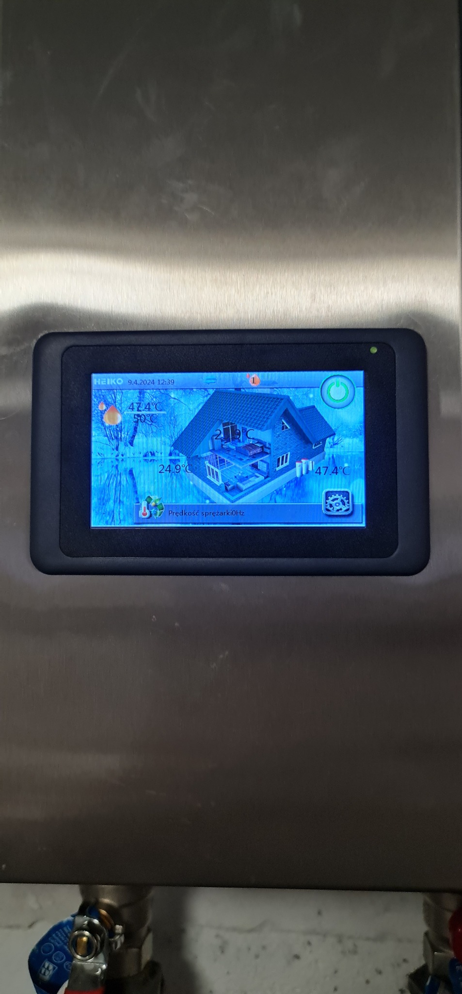 Ekran dotykowy panelu sterowania systemu grzewczego, wyświetlający schemat domu z temperaturami i parametrami pracy, zamontowany na metalowej obudowie.